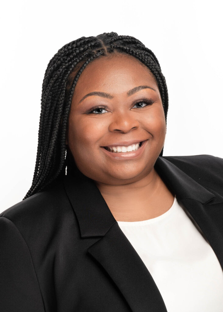 A woman with long braided hair smiles at the camera in a professional business headshot. She wears a black blazer over a white top against a plain white background, showcasing personal branding photography by a Durham NC photographer.