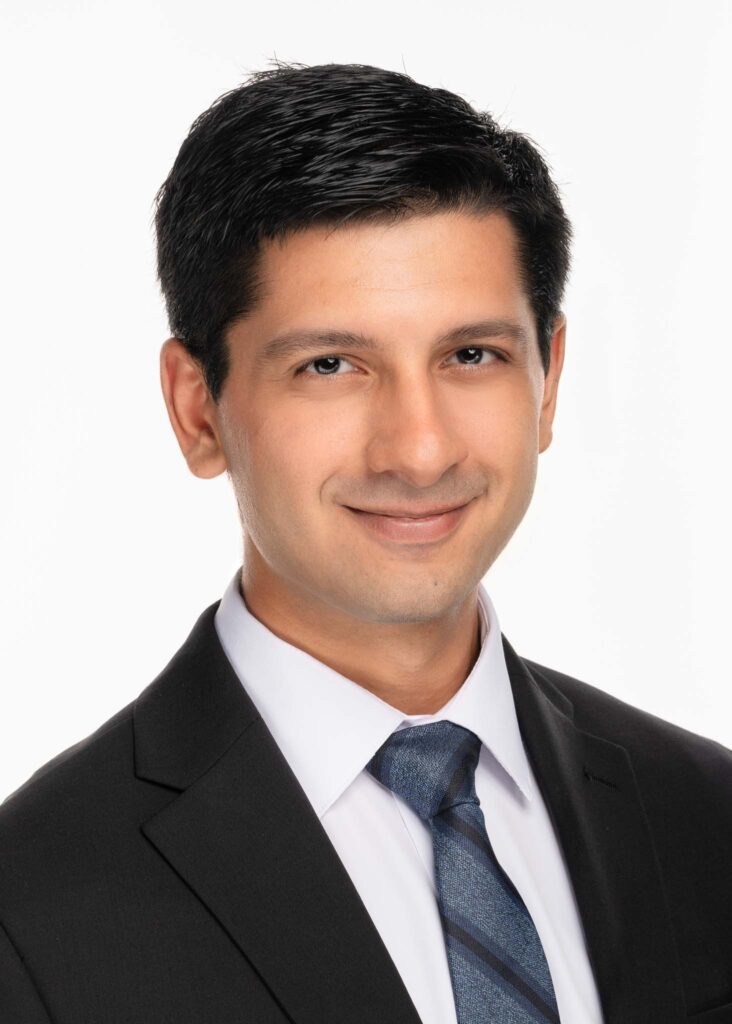 A man with short dark hair, dressed in a black suit, white shirt, and blue striped tie, smiles softly against a plain white background&mdash;ideal for personal branding photography or professional business headshots.