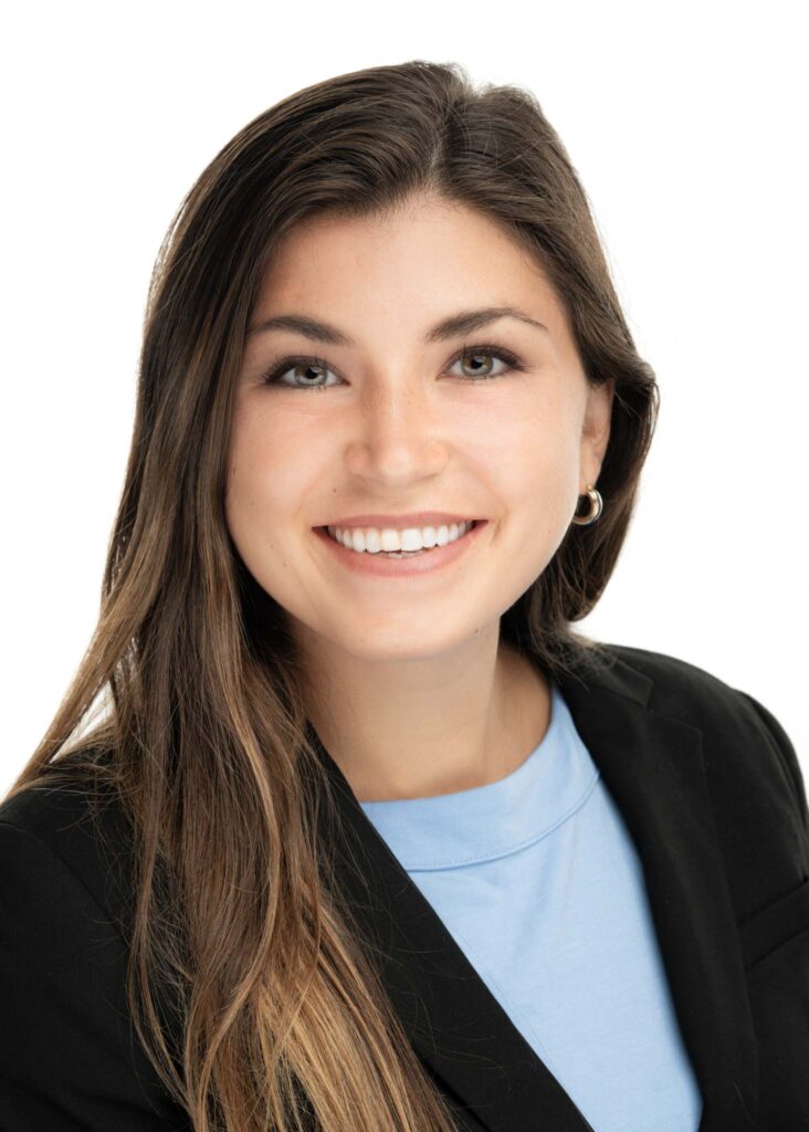 A young woman with long brown hair, wearing a black blazer over a light blue top, smiles at the camera against a plain white background&mdash;an ideal look for professional headshots Durham or actor headshots.