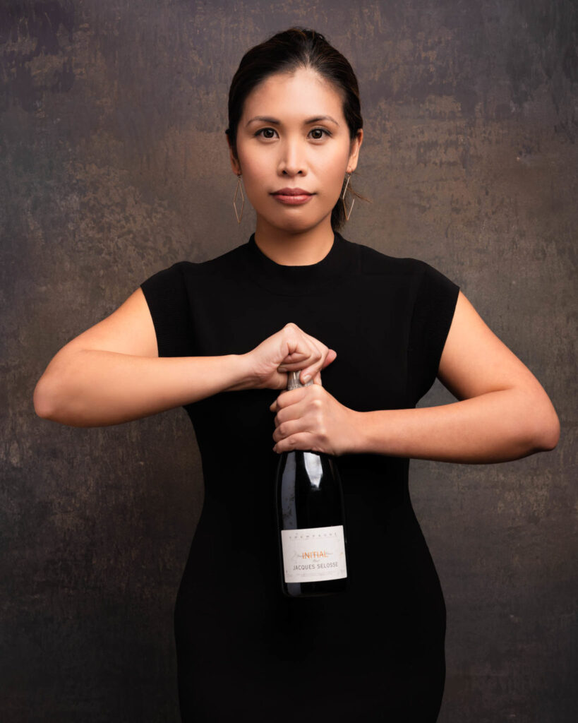 A woman in a black dress stands against a textured brown background, holding a bottle of sparkling wine upright with both hands. She looks directly at the camera with a neutral expression, perfect for personal branding photography or business headshots.