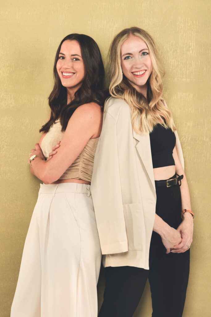 Two women stand back-to-back, smiling at the camera against a yellow background—an ideal example of personal branding photography by a Durham NC photographer. One has brown hair in beige attire; the other sports blonde hair with a beige jacket over black.