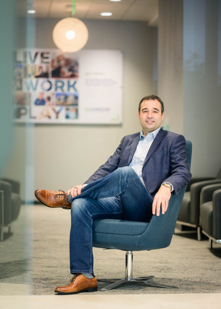 A man in a blue blazer, checked shirt, and jeans sits confidently in a modern office chair with his legs crossed. The setting, ideal for personal branding photography or business headshots, features a motivational LIVE WORK PLAY sign.