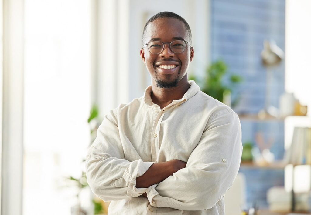 A smiling man wearing glasses and a light-colored shirt stands with his arms crossed in a bright, modern room with plants and shelves&mdash;an ideal setting for professional headshots Durham or personal branding photography.