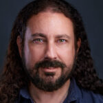A man with long curly dark hair, a beard, and mustache looks directly at the camera against a dark background. He is wearing a dark shirt and has a slight smile, perfect for personal branding photography or business headshots.