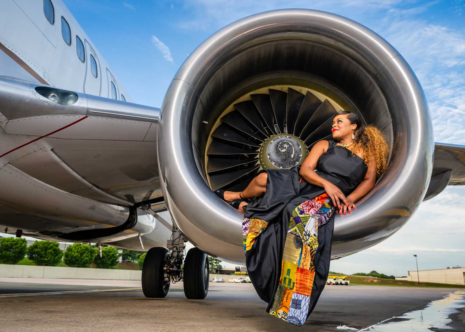 A woman in a long, colorful dress sits relaxed inside a jet engine on an airport tarmac, smiling and looking to the side under a blue sky&mdash;captured by a Durham NC photographer specializing in personal branding photography.