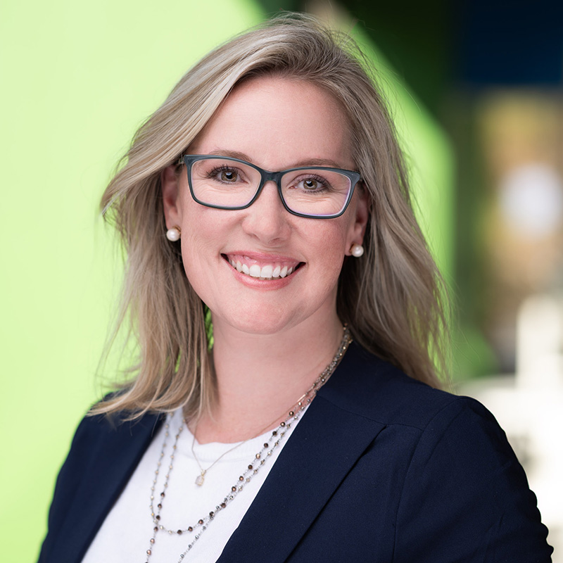 A smiling woman with blonde hair, glasses, a navy blazer, and layered necklaces stands in front of a blurred green and blue background&mdash;an ideal example of professional headshots Durham by a skilled Durham NC photographer.