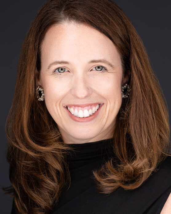 A woman with long brown hair, wearing a black top and statement earrings, smiles warmly at the camera against a dark background&mdash;captured by a Durham NC photographer specializing in professional headshots.