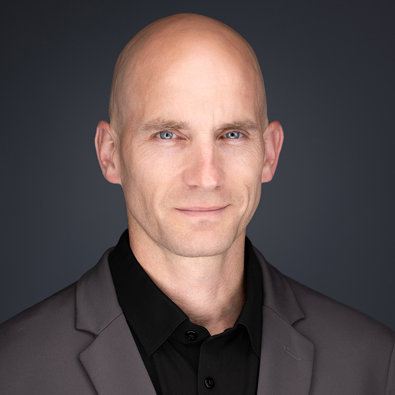 A bald man with light skin, blue eyes, and a slight smile is wearing a dark gray suit jacket over a black collared shirt, posing for business headshots against a plain dark background.
