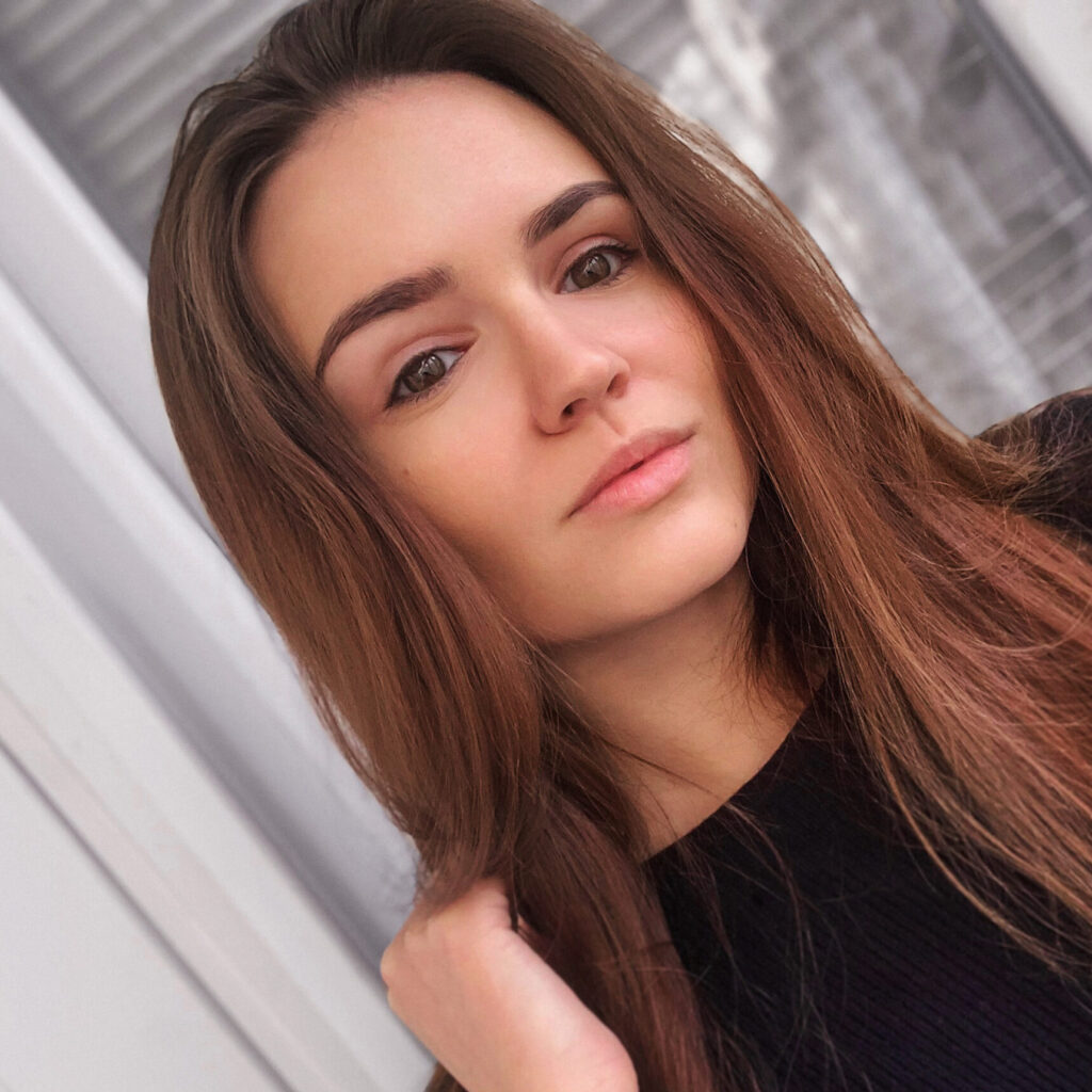 A young woman with long brown hair and a black top looks at the camera with a neutral expression. She stands near a window with white frames, captured by a Durham NC photographer in natural indoor light.