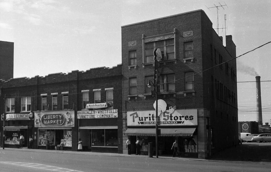 Black-and-white photo of a city street with several storefronts, including Liberty Market and a lunch counter. A few people stand on the sidewalk. Captured by a Durham NC photographer, smoke stacks are visible in the background.