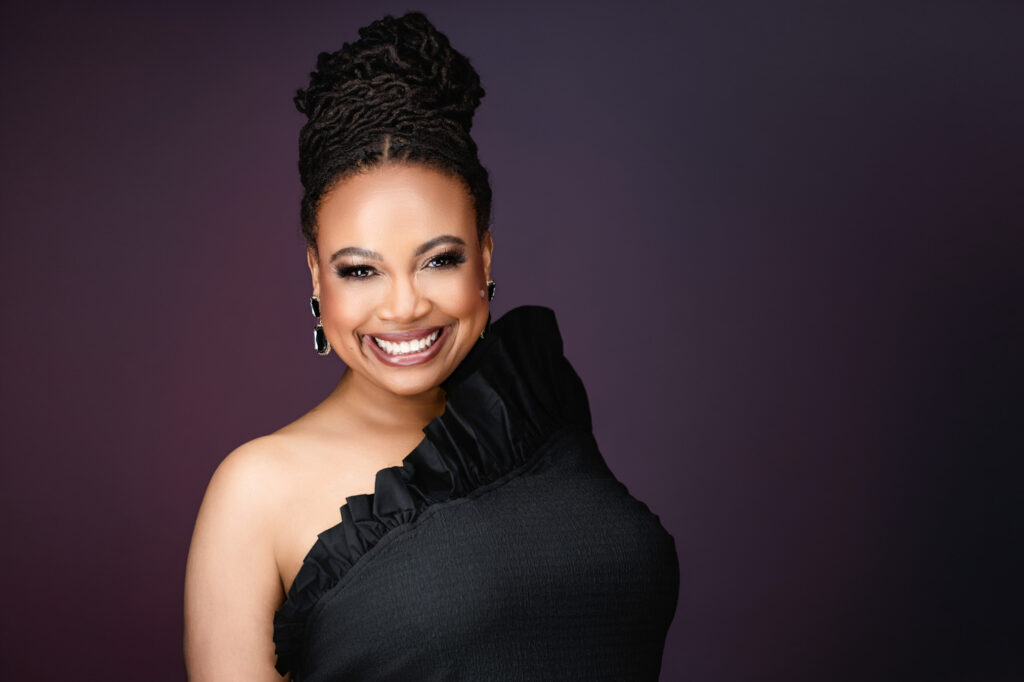 A woman with dark braided hair in an updo smiles, wearing a one-shoulder black dress with ruffle detail and dangling earrings, posed against a dark purple background, captured by a modeling photographer Durham.