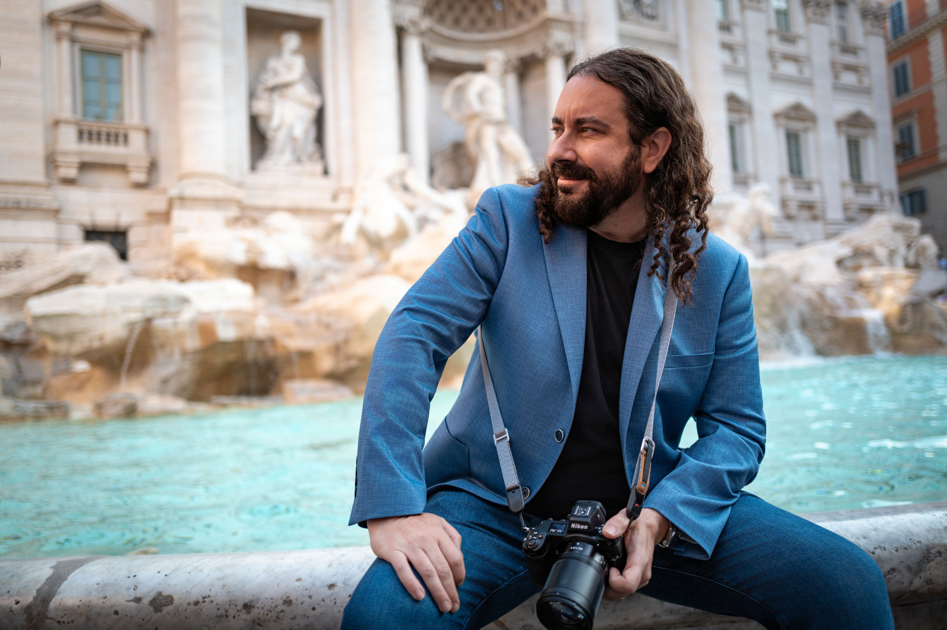A man with long curly hair and a beard, wearing a blue blazer and black shirt, sits near the edge of a fountain holding a camera. Captured by a modeling photographer Durham, historic stone architecture forms the backdrop.