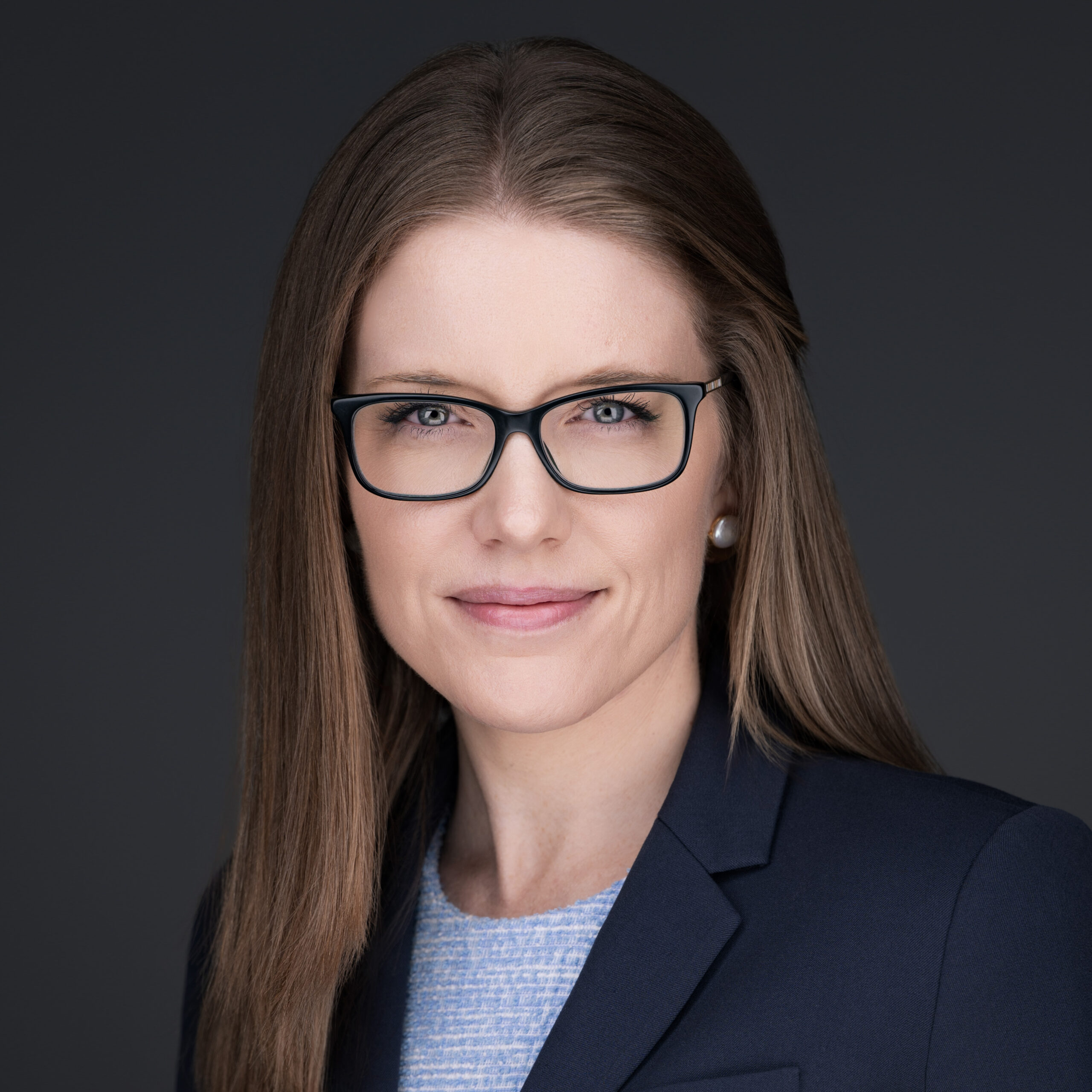 Professional headshot by a Durham NC photographer of a woman with straight brown hair, glasses, navy blazer, and light blue top, posing confidently against a dark gray background with a slight smile.